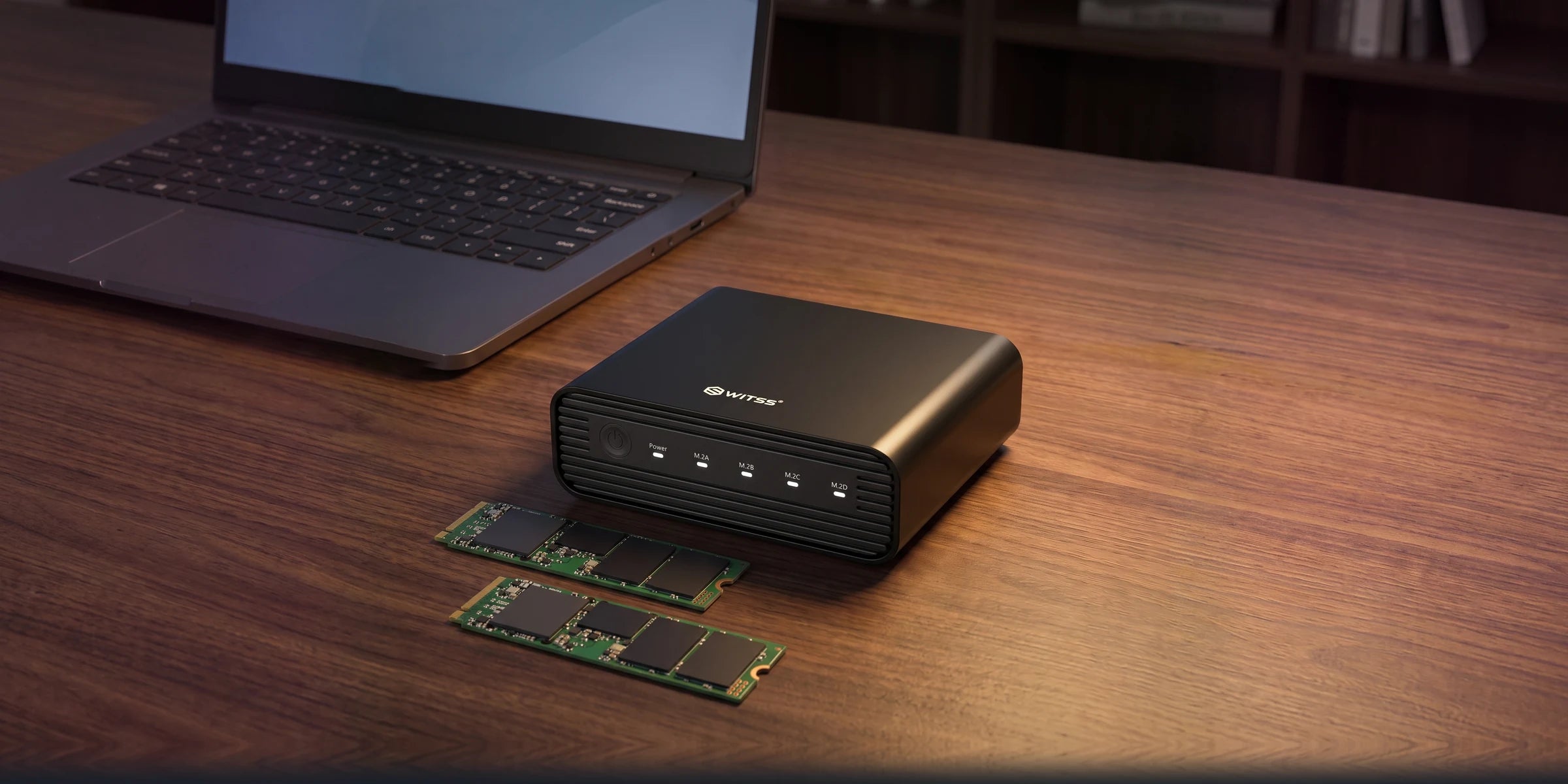 De Switss 4-Bay M.2 behuizing op een houten bureau, met twee losse M.2 NVMe SSD’s ervoor en een laptop op de achtergrond