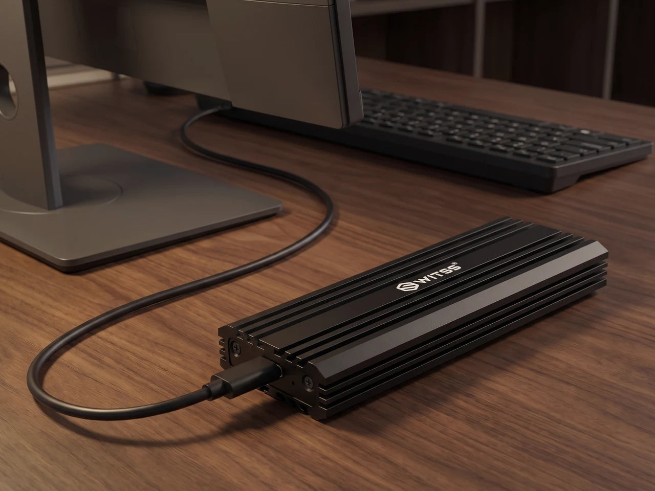 The Switss M.2 enclosure with its USB-C cable on a wooden desk, next to a monitor and a keyboard